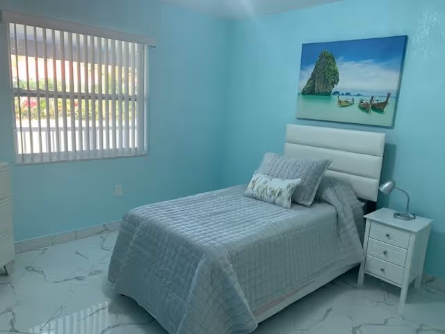 A neatly made single bed with blue-gray bedding and a white headboard in a light turquoise bedroom with a window, nightstand, and beach painting on the wall.