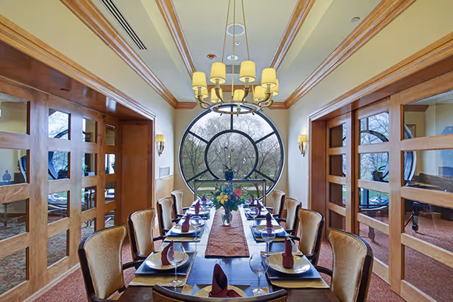 A formal dining room with a long rectangular table set for eight people. The table has a brown runner, plates, wine glasses, and folded napkins. The room features wooden framed glass doors on both sides and a large circular window at the far end with a view of trees outside. A chandelier with multiple lamps hangs above the table.