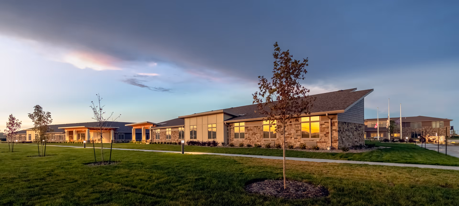 Exterior view of a senior living facility named Brookestone Gardens during sunset, showing a long, single-story building with stone and siding facade, several windows reflecting the sunset, a well-maintained lawn with young trees, and a paved walkway.