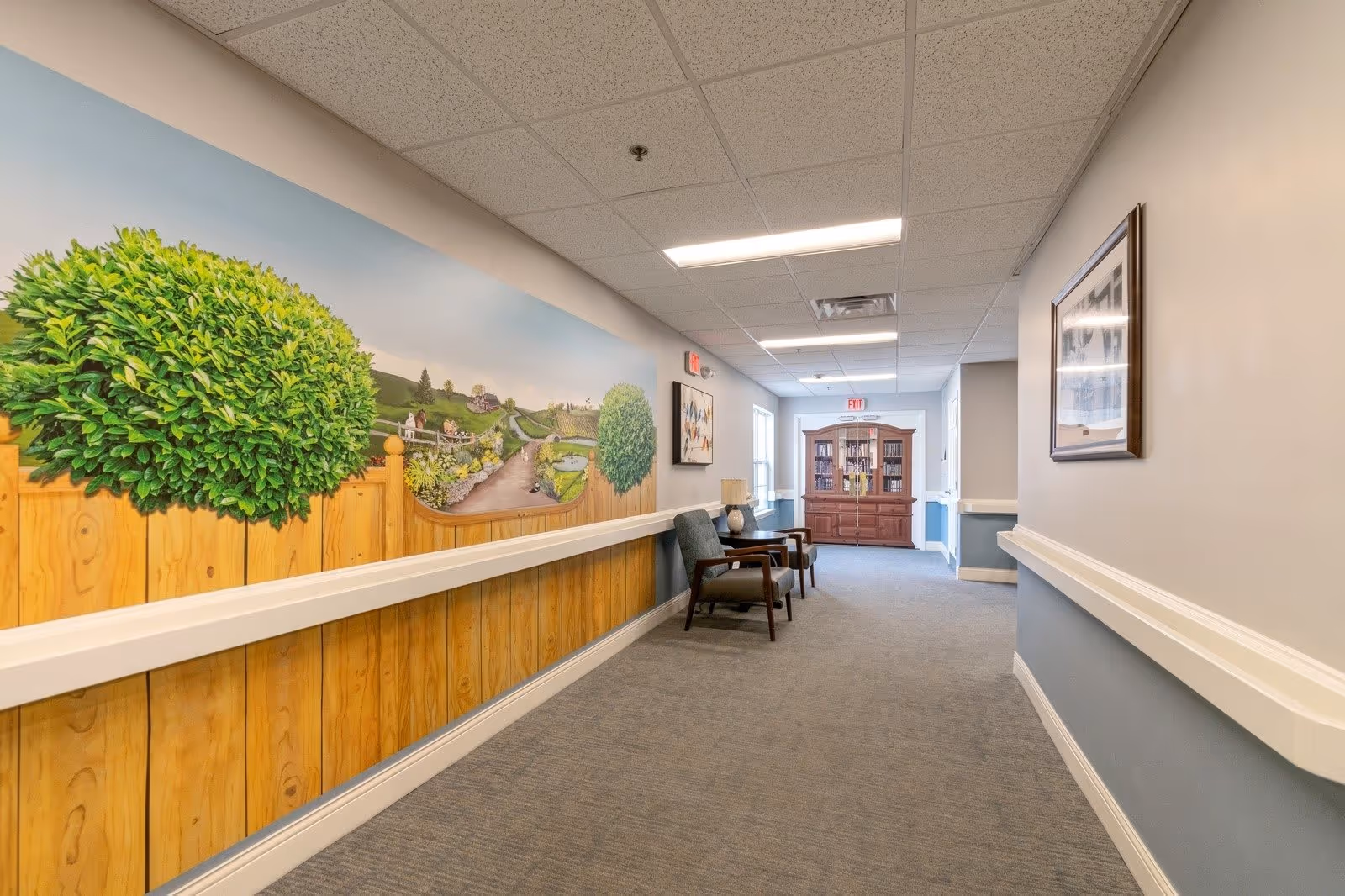 A well-lit hallway in a senior living facility with carpeted floor and light-colored walls. On the left wall, there is a mural depicting a wooden fence with green bushes and a scenic countryside landscape. Two chairs and a small table with a lamp are placed along the right side of the hallway. At the end of the hallway, there is a wooden cabinet with glass doors containing books or decorative items. The ceiling has recessed lighting panels.