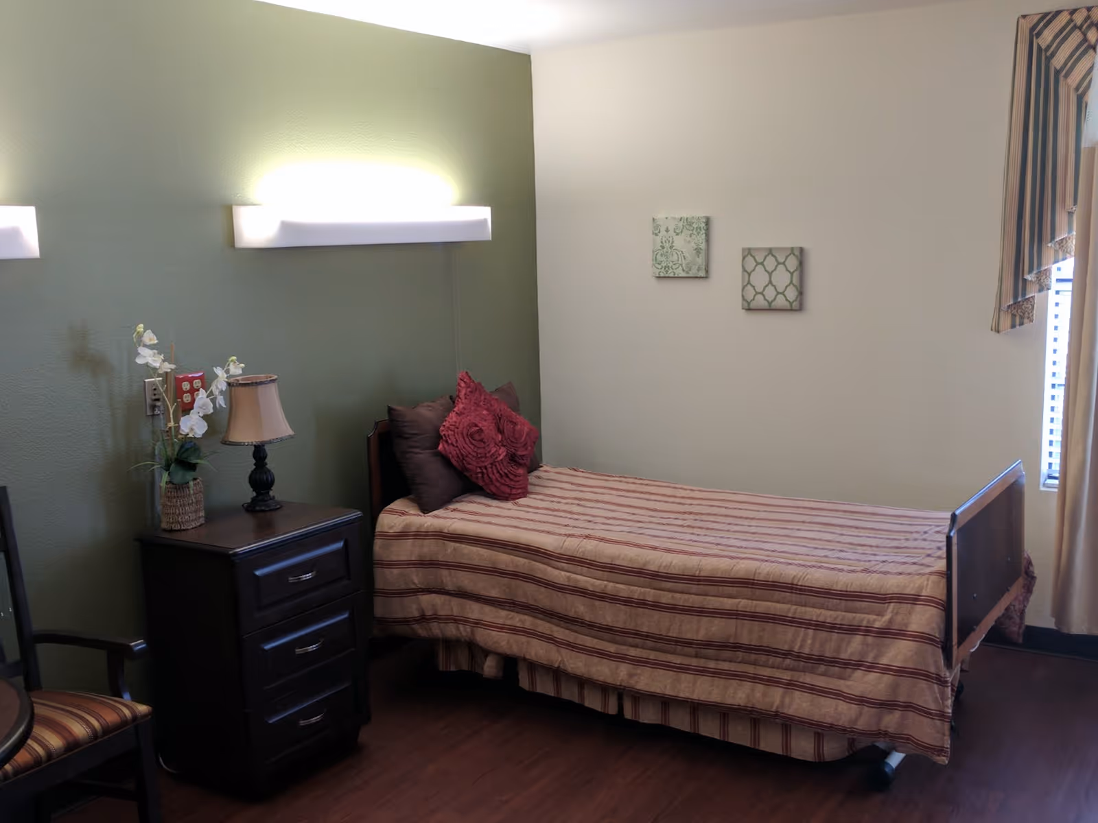 A single bed with striped bedding and decorative pillows in a room with green and white walls. Next to the bed is a dark wooden nightstand with a lamp and a vase with white flowers. There is a chair with striped upholstery and two small framed artworks on the wall. A window with striped curtains is partially visible on the right.