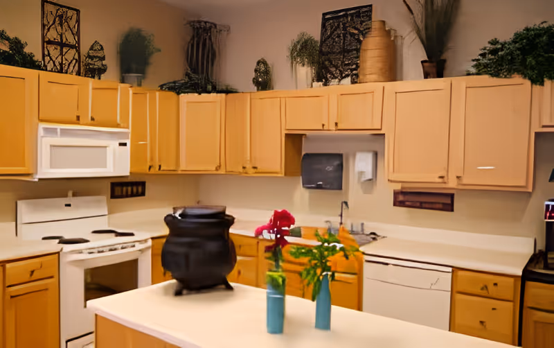 A kitchen with light wood cabinets, a white stove, microwave, and dishwasher. There is a central island with a black pot and two small vases holding flowers. Decorative plants and artwork are placed above the cabinets.