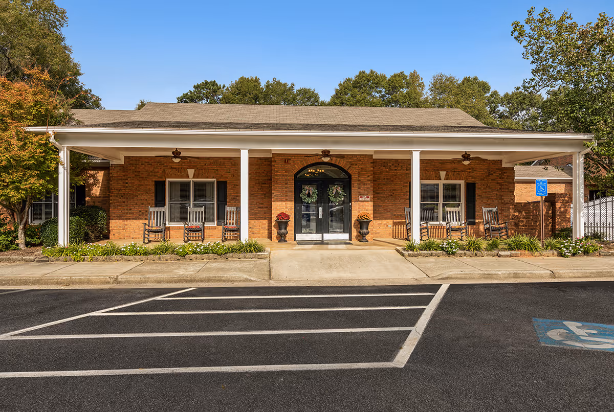 Front exterior view of a single-story brick building with a covered porch featuring several rocking chairs and two large planters with flowers on either side of a double glass door entrance. There are trees and shrubs around the building, and a parking area with a handicapped parking space in front.