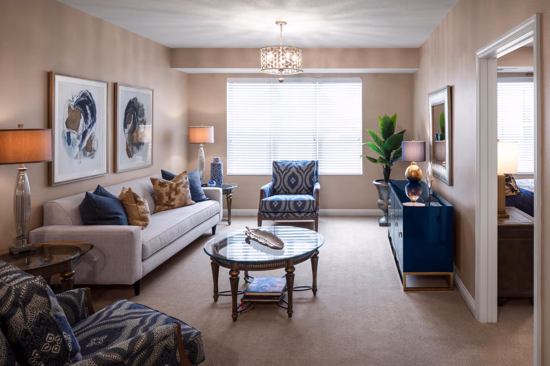 Bright furnished living room with a sofa, patterned armchairs, a glass coffee table, sideboard, lamps and a window with blinds.