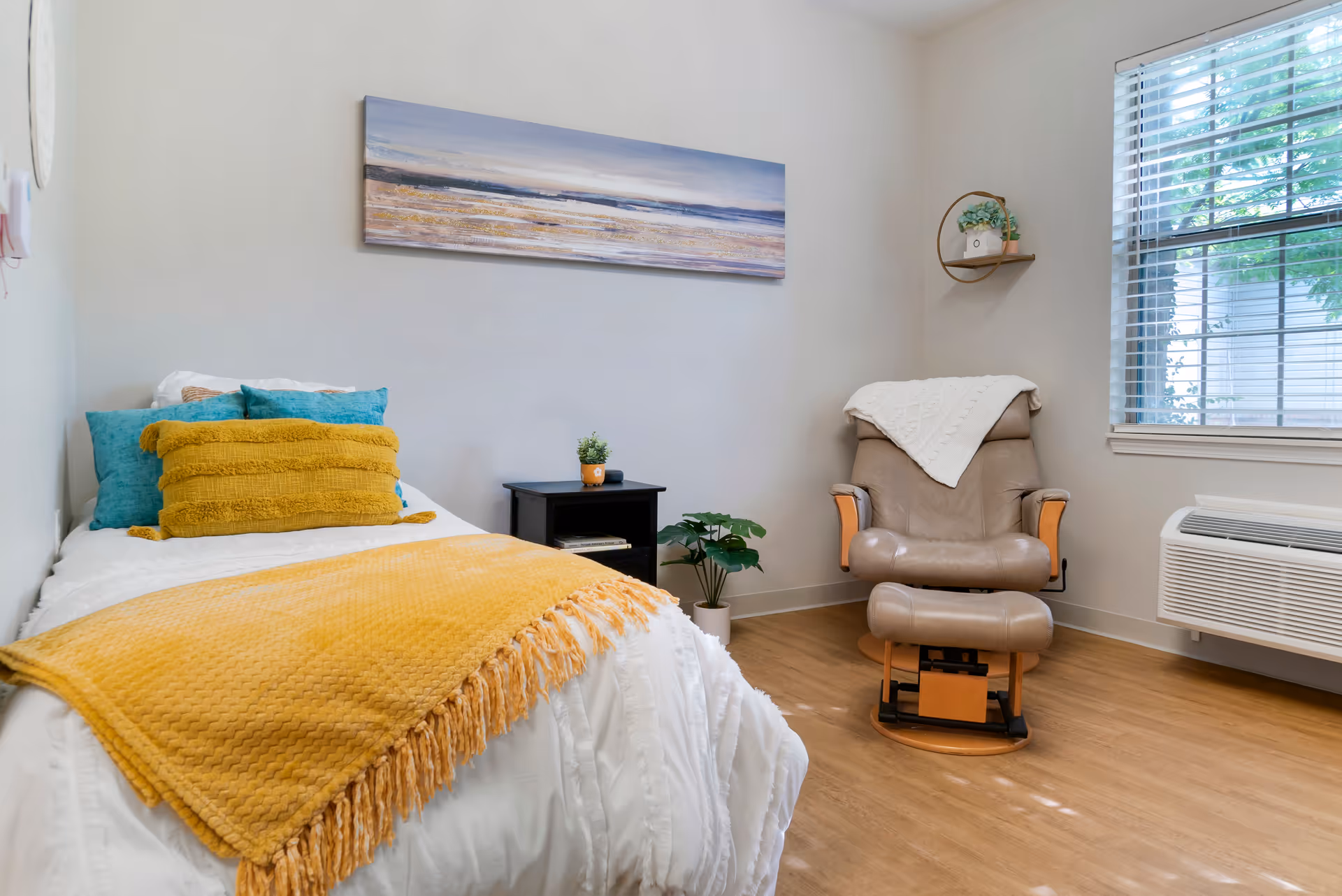 A cozy bedroom with a single bed covered in white bedding and a mustard yellow throw blanket and pillows. Next to the bed is a small black nightstand with a small plant on it. Across from the bed is a comfortable beige recliner chair with a white knitted throw draped over the back. A window with blinds lets in natural light, and a small round shelf with a plant is mounted on the wall. The floor is wooden, and a landscape painting hangs above the bed.