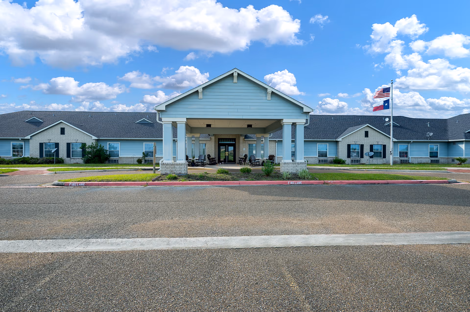 Front exterior view of a single-story senior living facility building with a covered entrance supported by white columns. The building has light blue siding with white brick accents and a dark shingled roof. There are American and Texas flags on a flagpole to the right of the entrance. The sky is blue with scattered clouds.