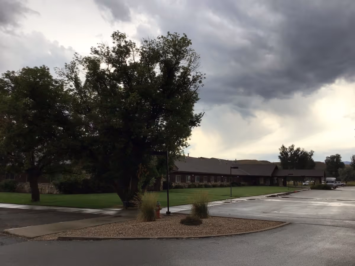 Exterior view of Westview Health Care Center showing a long, single-story building with a covered entrance, surrounded by green lawns, trees, and a wet driveway under a cloudy sky.
