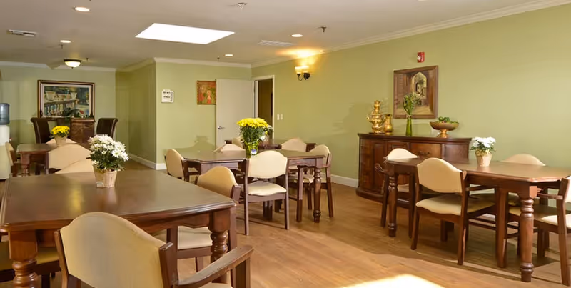 A dining room with several wooden tables and beige cushioned chairs. Each table has a small flower arrangement. The walls are painted light green and decorated with framed artwork. There is a wooden sideboard with decorative items and a vase with flowers. The floor is wooden, and the room is well-lit with ceiling lights and wall sconces.