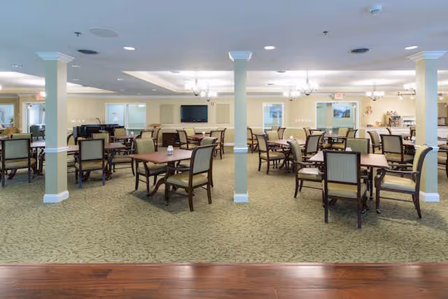 Spacious dining room with multiple wooden tables and cushioned chairs arranged neatly on a patterned carpet. The room has beige walls with white columns and ceiling lights, and a television mounted on the far wall.