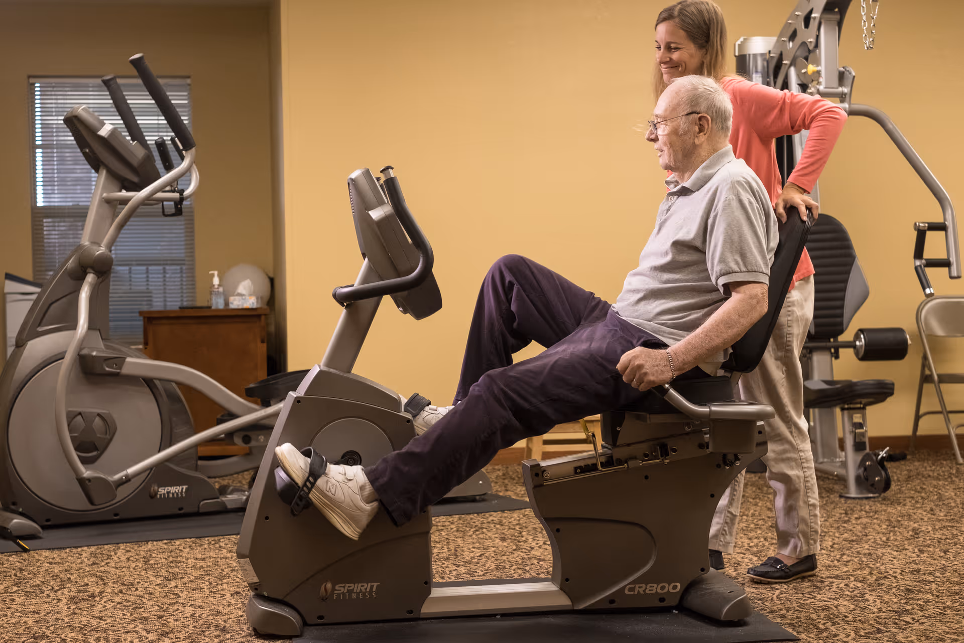 An elderly man exercises on a recumbent stationary bike in a fitness room while a woman stands behind him, offering support and encouragement. The room has exercise equipment including an elliptical machine and weight machines, with beige walls and carpeted floor.