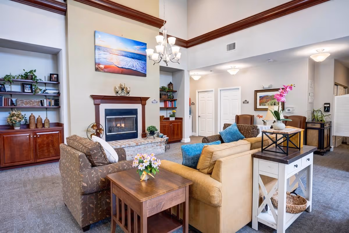 A cozy living room area in a senior living facility featuring a fireplace with a beach-themed painting above it, two sofas with cushions, a wooden side table with a flower vase, built-in shelves with books and decorative items, and a chandelier hanging from a high ceiling.
