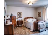 A cozy bedroom with a wooden bed frame, white bedding, and a quilt draped over the foot of the bed. The room features vintage wooden furniture including a dresser and a nightstand with a lamp. Two framed portraits hang on the walls, and a window with light curtains allows natural light to fill the space.