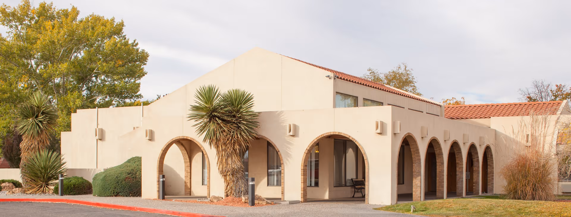 Low-rise stucco building with arched colonnade, desert plants, and a red-tiled roof under a cloudy sky.