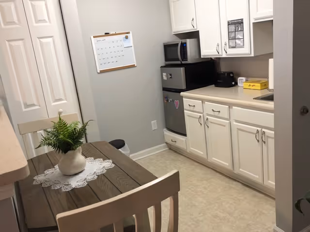 Small kitchen area with white cabinets, a microwave, mini refrigerator, toaster, and a sink. A wooden table with two chairs is in the foreground, decorated with a white doily and a potted green plant. A wall calendar is hanging on the wall near a closed door.