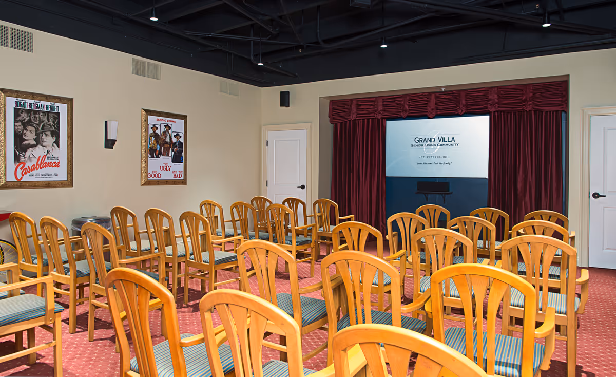 Rows of wooden chairs face a small stage and projection screen framed by red curtains in a senior living community screening room.