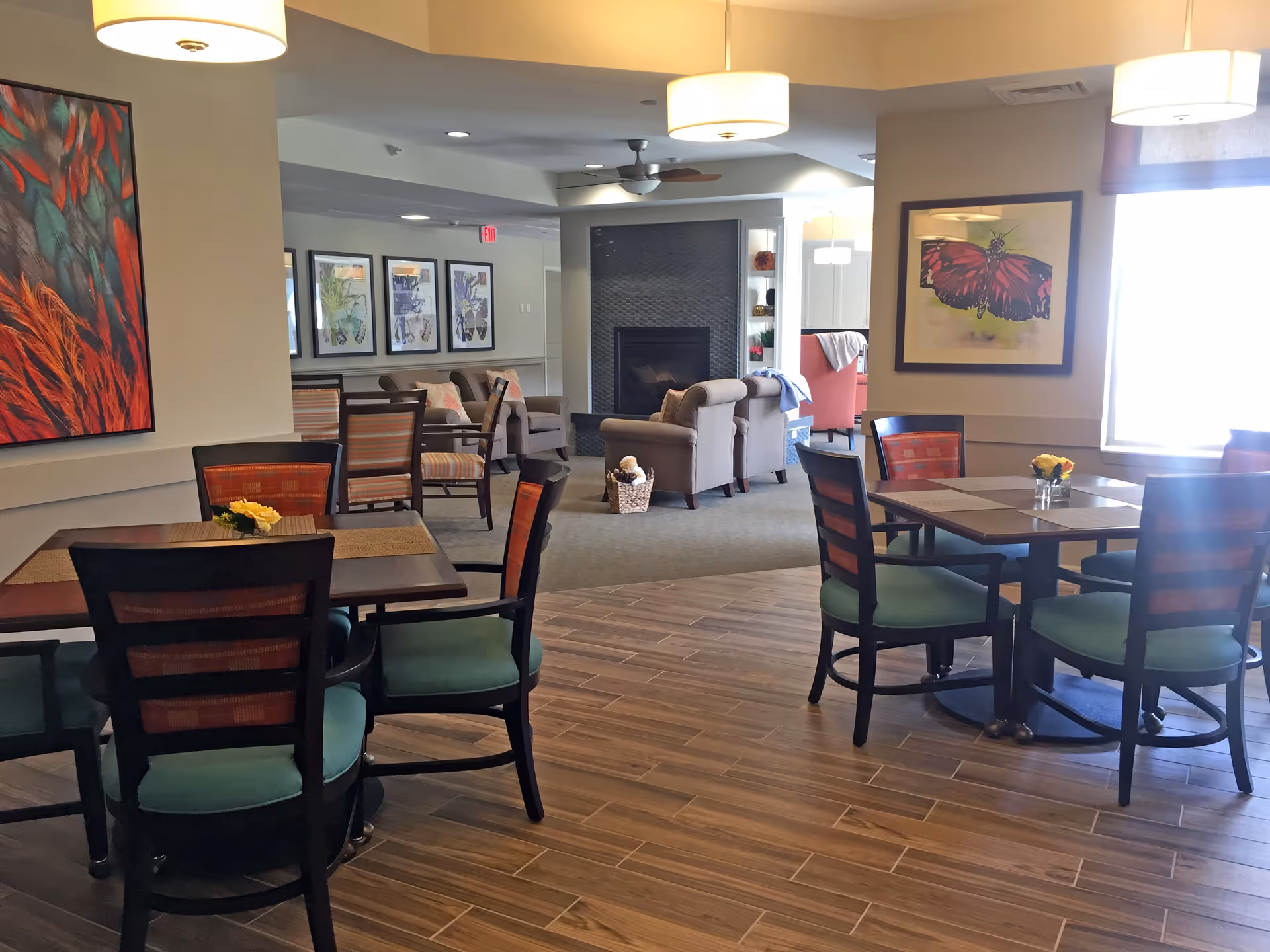 Interior view of a senior living facility common area with dining tables and chairs in the foreground and a seating area with armchairs and a fireplace in the background. The room is decorated with colorful artwork on the walls and has pendant lights hanging from the ceiling.