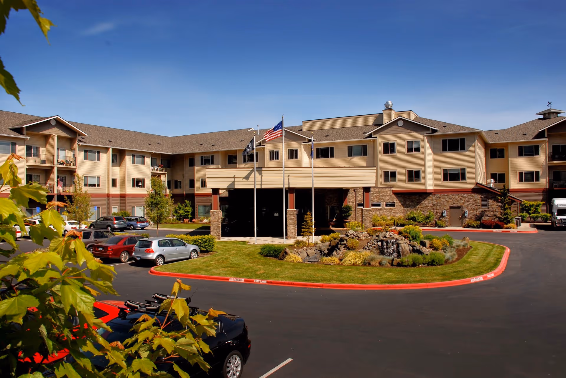 Three-story beige senior living building with a covered entrance, flagpoles, parked cars, and a landscaped circular driveway.