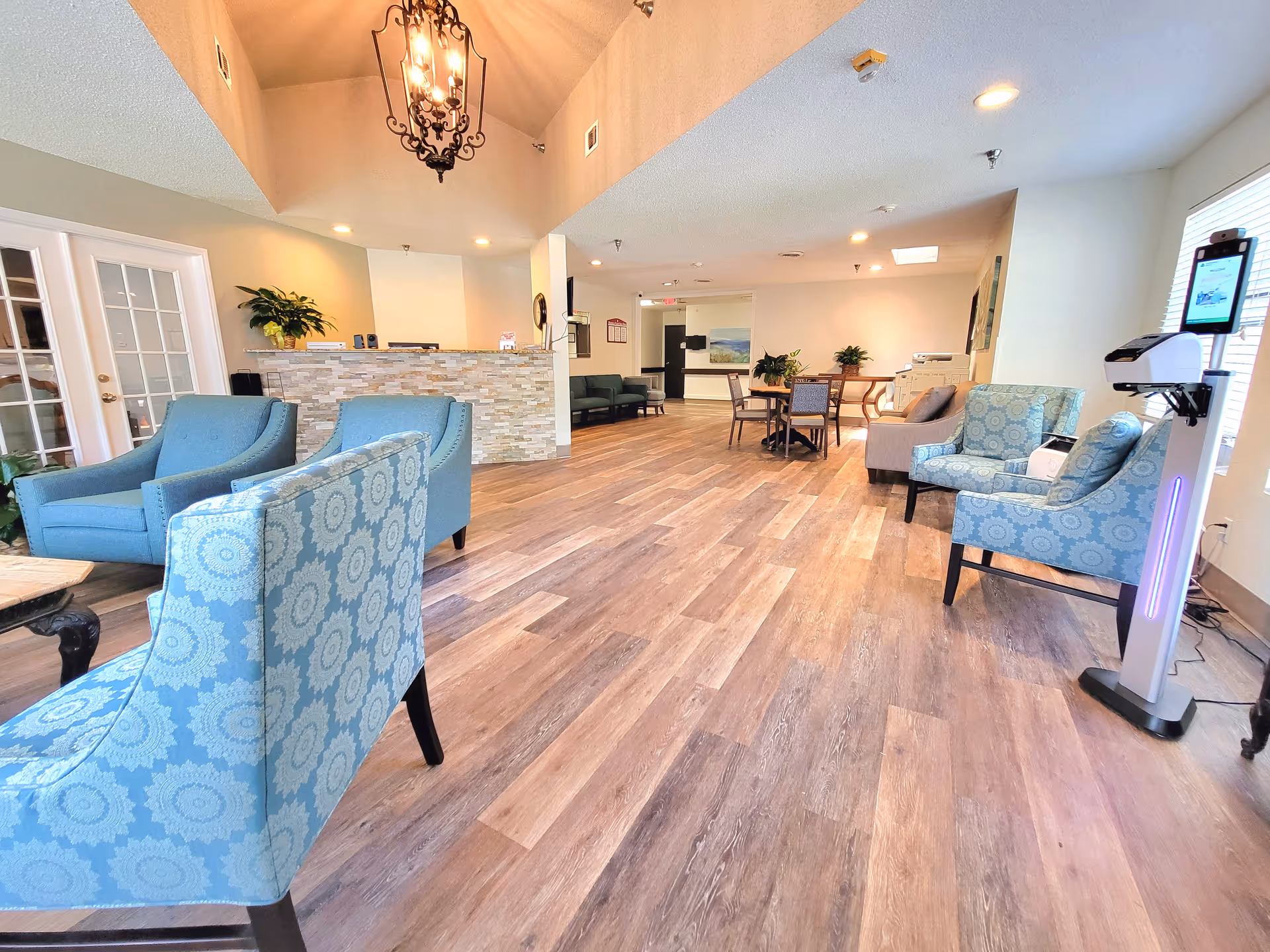 A spacious and well-lit reception area with wooden flooring, featuring a stone-faced reception desk, several blue and patterned armchairs, a round table with chairs, potted plants, and a hanging chandelier. There is a hand sanitizer station near a window on the right side.