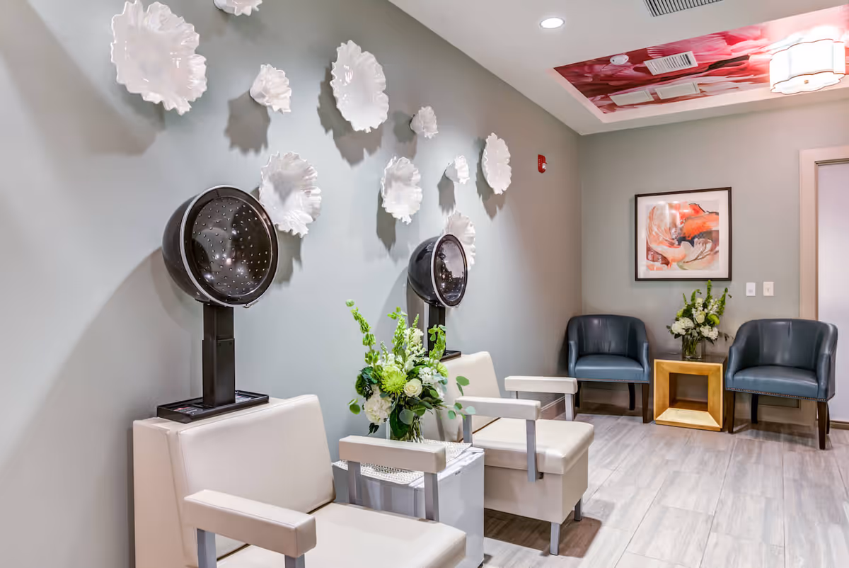 A modern salon area with two white salon chairs equipped with black hair dryers. The wall behind the chairs is decorated with white flower-shaped wall art. There is a small table with a floral arrangement between the chairs. In the background, there are two blue armchairs with a wooden side table holding another floral arrangement. The ceiling has a decorative panel with a pink floral design.