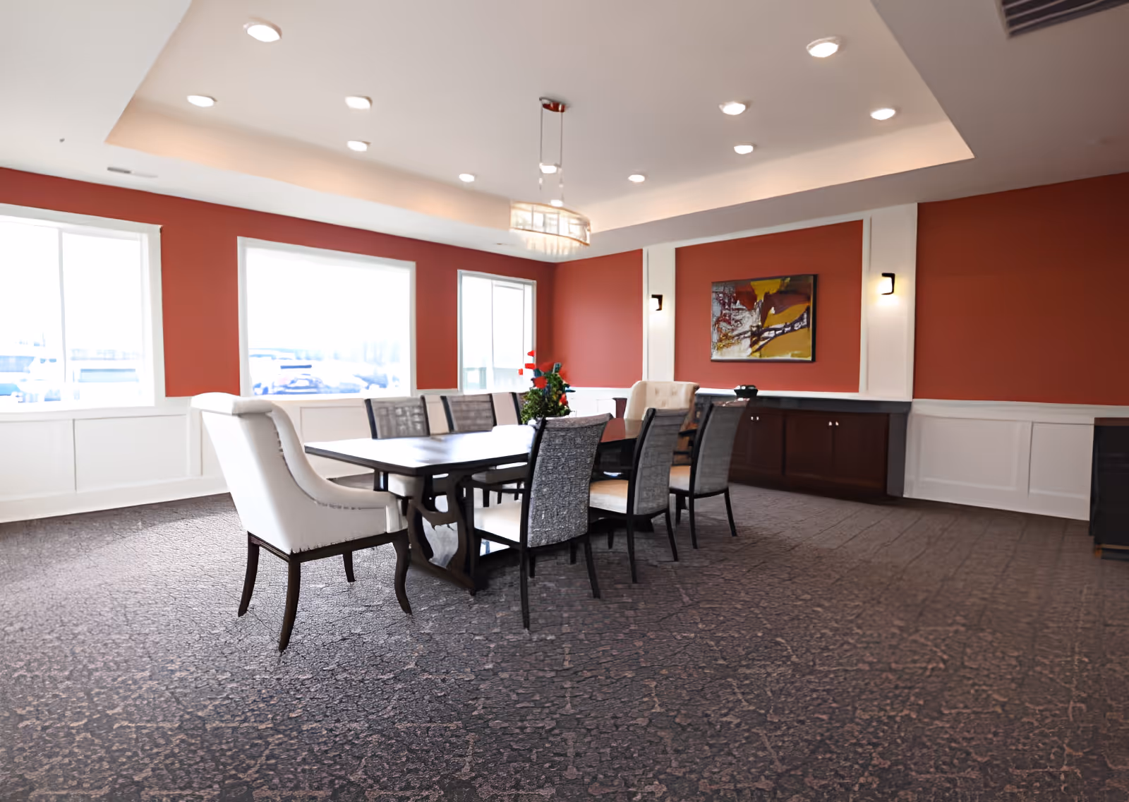 A dining room with a large rectangular wooden table surrounded by eight chairs, two of which have white upholstery and the others have patterned fabric. The room has red walls with white wainscoting, three large windows letting in natural light, a modern chandelier hanging from the ceiling, and a colorful abstract painting on the wall above a dark wooden cabinet.