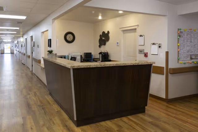 Reception desk area inside a senior living facility with a long hallway extending to the left. The desk has a granite countertop and dark wood paneling. Behind the desk are office supplies, a clock on the wall, and decorative wall art. The floor is wood, and the walls are light-colored with handrails along the hallway.