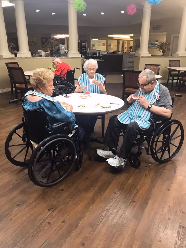 Three elderly individuals in wheelchairs sitting around a round table in a communal dining area, eating sandwiches. The room has wooden flooring, white columns, and colorful hanging decorations.