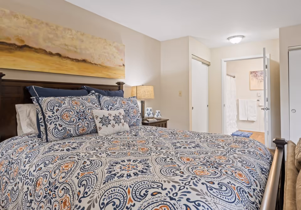 A bedroom with a large bed covered in a patterned blue, white, and orange comforter and matching pillows. There is a wooden headboard, a bedside table with a lamp and decorative items, and a large abstract painting above the bed. An open door reveals a bathroom with a shower curtain, towels, and a piece of artwork on the wall.