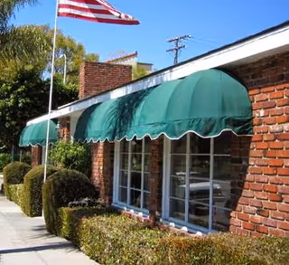 Exterior view of a brick building with green awnings over the windows, a neatly trimmed hedge along the sidewalk, and an American flag on a flagpole in front of the building under a clear blue sky.