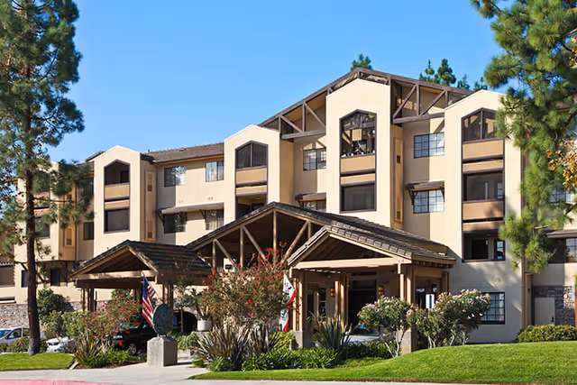 Exterior view of a multi-story senior living facility building with beige walls and brown trim, featuring a covered entrance with wooden beams, surrounded by landscaped greenery and trees under a clear blue sky.
