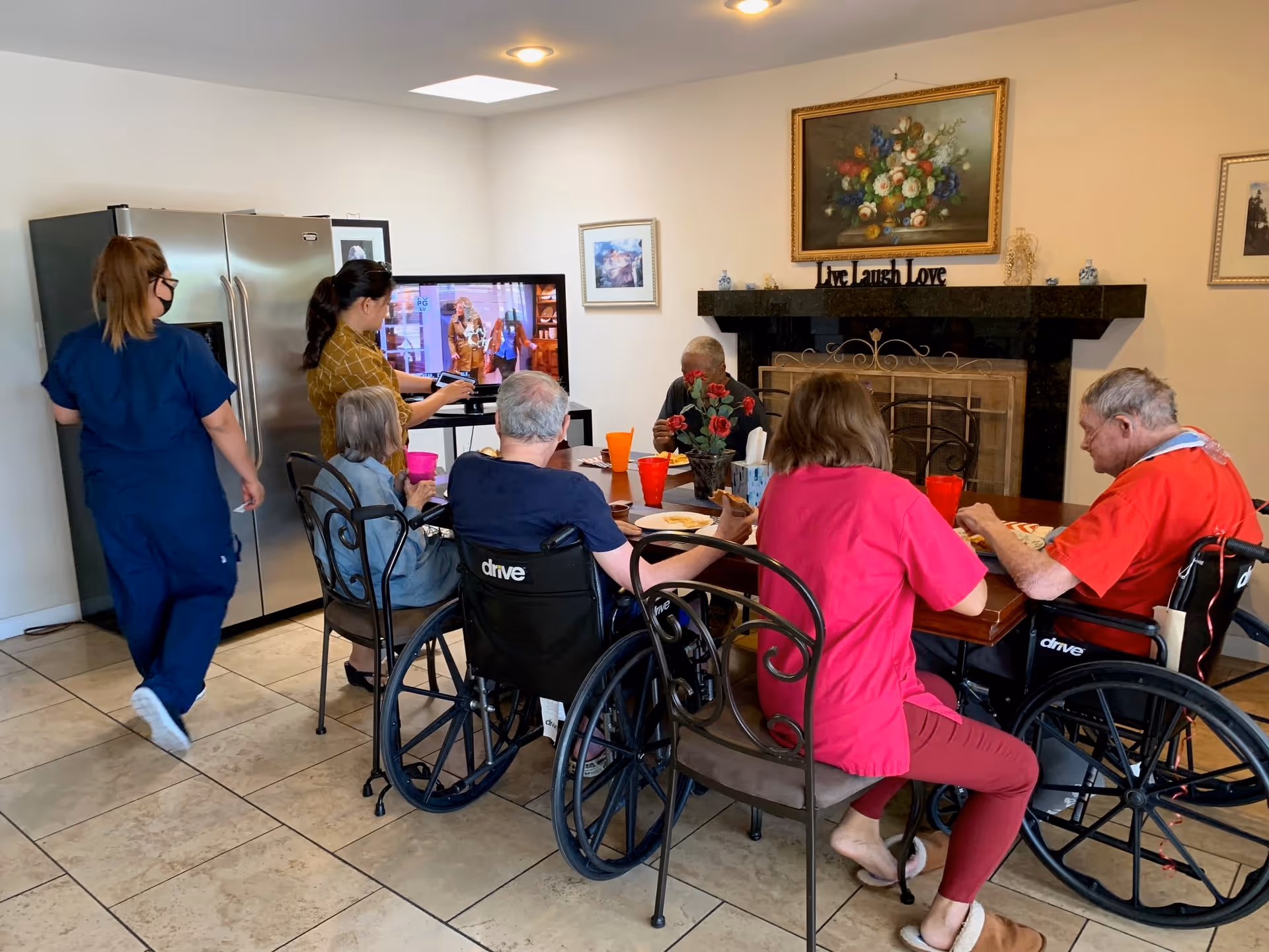 A group of elderly people, some in wheelchairs, sitting around a dining table eating and drinking. Two caregivers are present, one standing near a refrigerator and the other adjusting a television screen. The room has a fireplace with a floral painting above it and a sign that reads 'Live Laugh Love'.