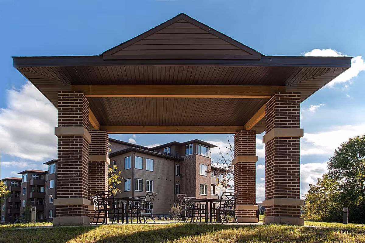 Outdoor covered seating area with brick pillars and a wooden roof, featuring several metal tables and chairs on a grassy area, with a multi-story residential building in the background under a partly cloudy blue sky.