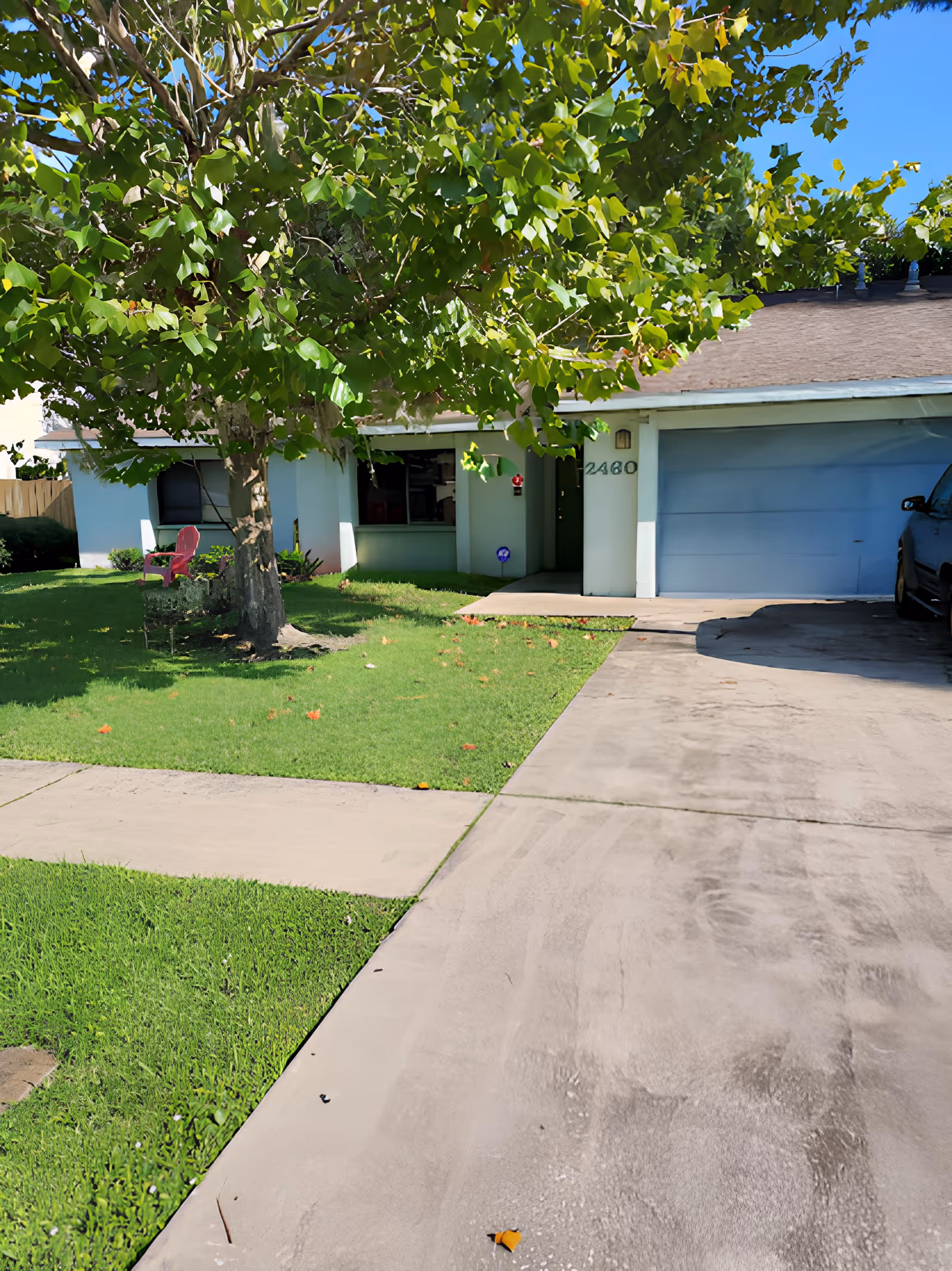 Front exterior view of a single-story house with a large tree in the front yard, green grass, a concrete driveway, and a garage. There is a red chair on the lawn near the house.