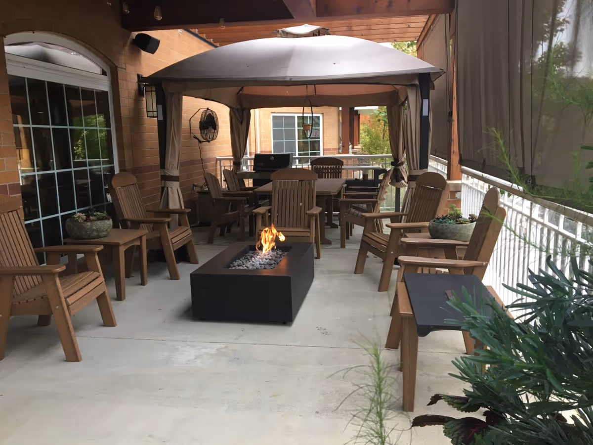 Outdoor patio area with wooden chairs arranged around a rectangular fire pit with flames. There is a canopy providing shade over a dining table and chairs in the background. Potted plants and greenery are visible along the edges of the patio, which is adjacent to a brick building.