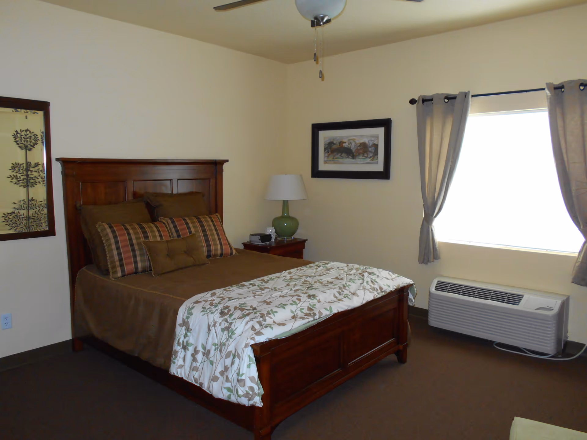A bedroom with a wooden bed frame and headboard, made up with brown and plaid pillows and a floral blanket. There is a nightstand with a green lamp and alarm clock next to the bed. A window with beige curtains and an air conditioning unit below it is on the right wall. The walls are painted light beige and there is framed artwork hanging above the nightstand and on the left wall.