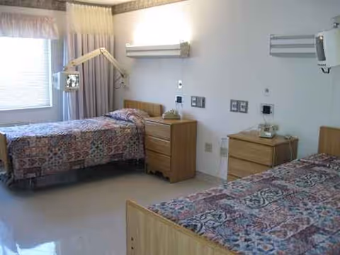 Two single beds in a bright room with nightstands, curtains, and medical equipment.
