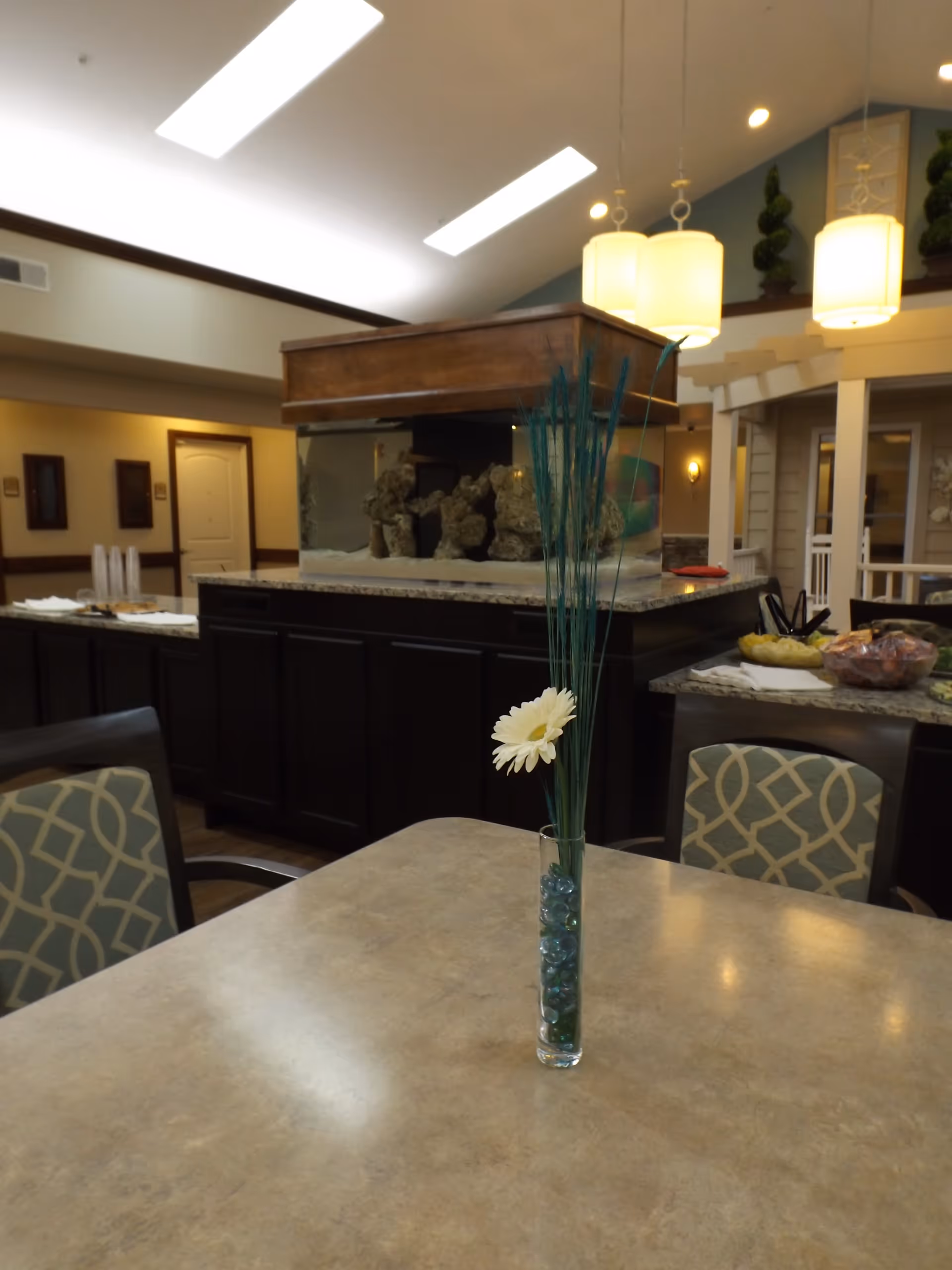 Interior view of a senior living facility common area with a table in the foreground featuring a tall glass vase containing a single white flower and decorative blue stones. Behind the table is a large aquarium with rocks inside, set on a dark wooden cabinet. The room has high ceilings with modern hanging lights and patterned chairs around the table. There are snacks and plates on a counter in the background.