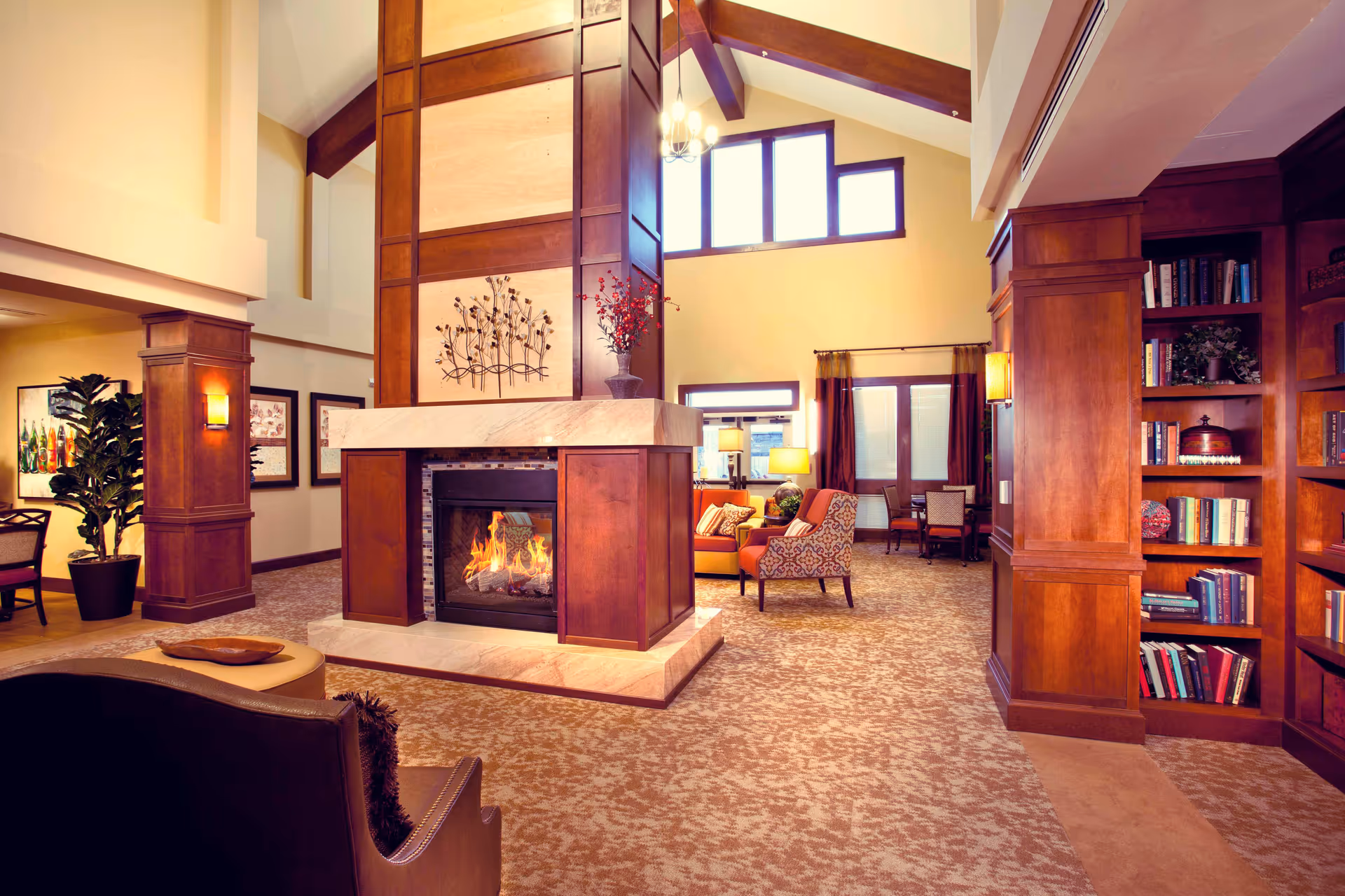 A spacious and warmly lit living room area featuring a central double-sided fireplace with wooden paneling and a decorative metal wall art above it. The room has high ceilings with large windows letting in natural light. There are comfortable chairs and sofas arranged around the fireplace, a bookshelf filled with books on the right, and framed artwork on the walls. The carpet has a patterned design, and there are plants and lamps adding to the cozy ambiance.