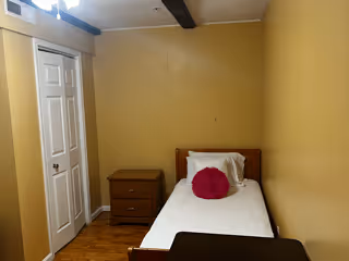 A small bedroom with a single bed against a mustard yellow wall. The bed has white bedding and a round pink pillow. Next to the bed is a wooden nightstand with two drawers. There is a white closet door on the left side of the room and a dark wooden beam on the ceiling.