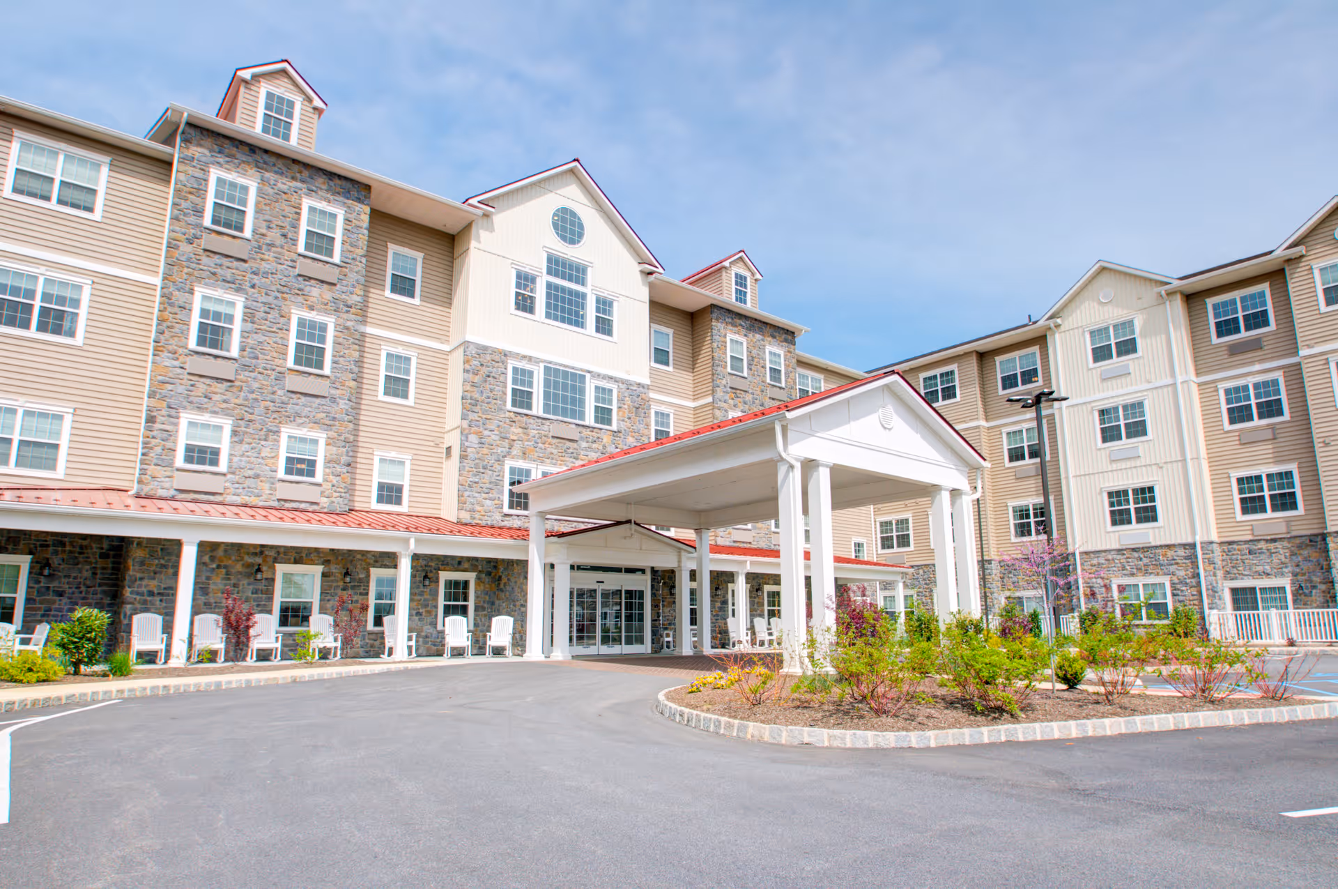 Front exterior view of a multi-story senior living facility with a covered entrance, stone and beige siding, multiple windows, and a landscaped roundabout with shrubs and small trees.