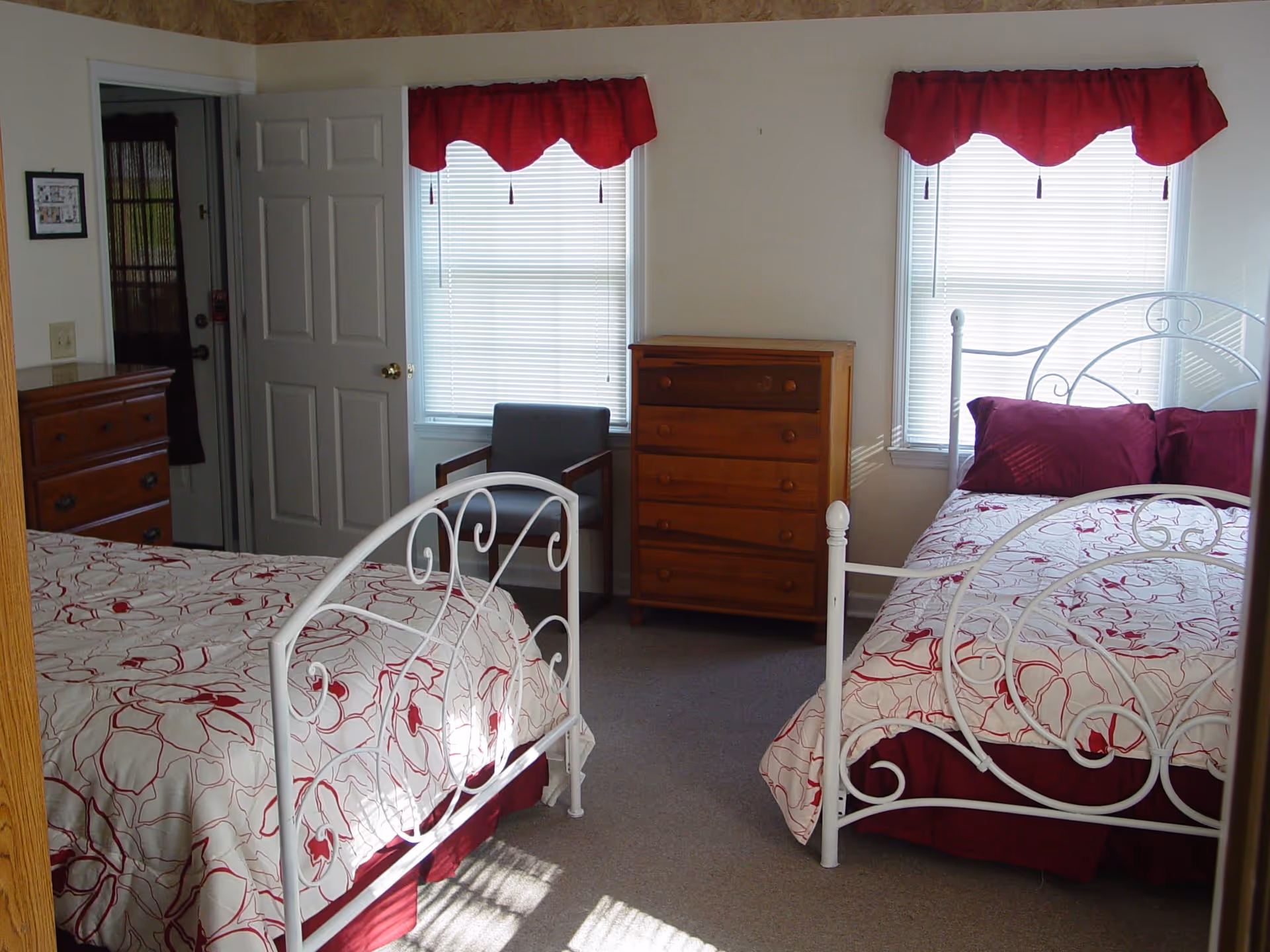 A bedroom with two white metal-framed beds, each with white and red floral bedding and red pillows. Between the beds is a wooden chest of drawers, and a gray armchair is positioned near two windows with red valances and white blinds. A door is open on the left side of the room, and another wooden dresser is partially visible.