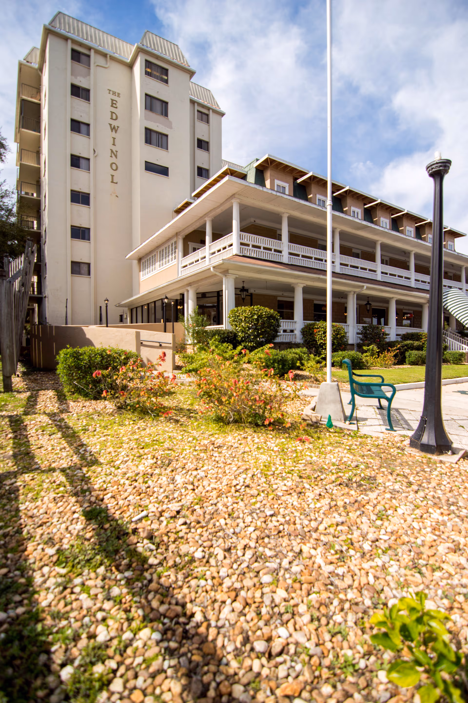 Exterior front of a multi-story senior living building with wraparound porches, landscaped gravel beds, and an outdoor bench.