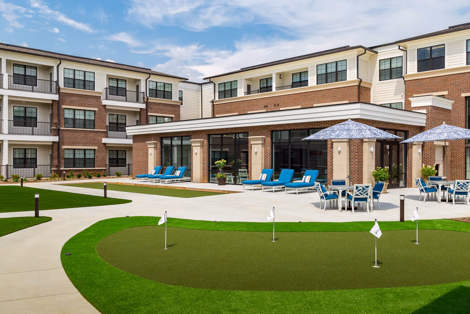Outdoor patio area of a senior living facility with lounge chairs, tables with umbrellas, and a small putting green in the foreground. The building has three stories with brick and siding exterior and large windows.