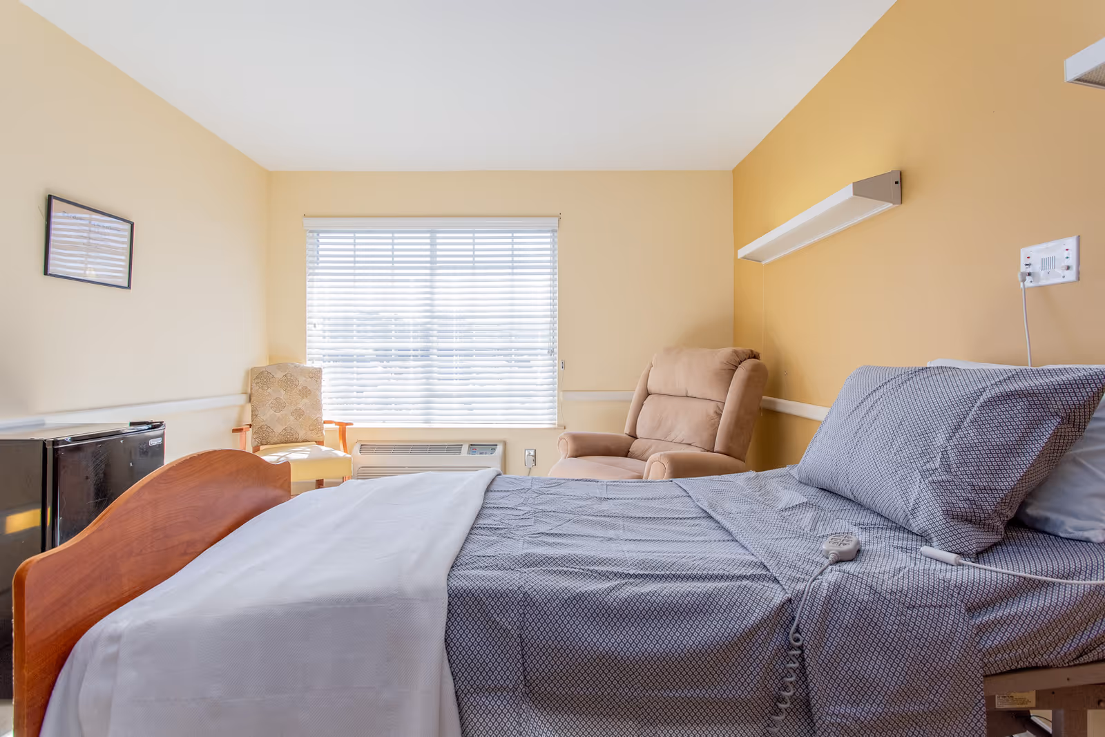 A bright nursing facility bedroom with a hospital-style bed made with gray patterned sheets and a white blanket. There is a beige recliner chair and a patterned armchair near a window with white blinds. A small black refrigerator is visible on the left side of the room, and the walls are painted in light yellow and beige tones.