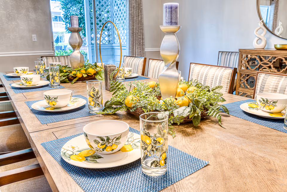 A dining table set with blue placemats, plates, bowls, and glasses decorated with lemon patterns. The table has a centerpiece with greenery, lemons, and two tall silver candle holders with purple candles. The room has striped upholstered chairs, a wooden sideboard with decorative items, and a window with curtains letting in natural light.