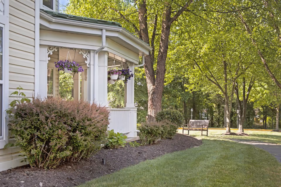 View of a porch with hanging flower pots and decorative trim on a light-colored building, surrounded by bushes and trees with a grassy lawn and a wooden bench in the background.