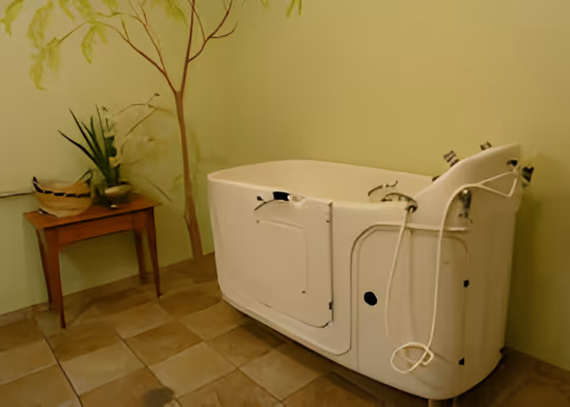A walk-in bathtub with safety handles and a door for easy access, placed against a light green wall. Next to it is a small wooden table with decorative plants and a bowl on top. The floor is tiled in a beige and brown pattern.