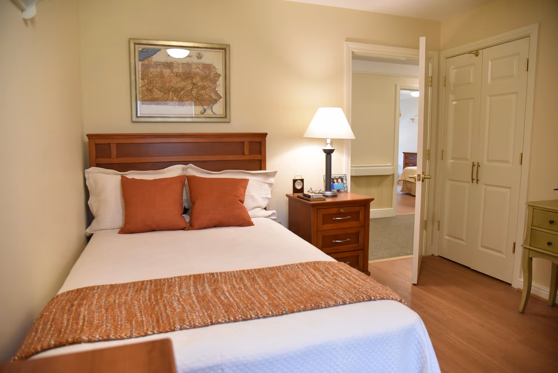 A cozy bedroom with a wooden headboard bed made with white bedding and two rust-colored pillows. A matching rust-colored throw blanket is folded at the foot of the bed. Next to the bed is a wooden nightstand with a lamp, a clock, and a framed photo. The room has beige walls, a framed map hanging above the bed, and an open door leading to another room with similar decor. There is a white double-door closet and a small green table on the right side.