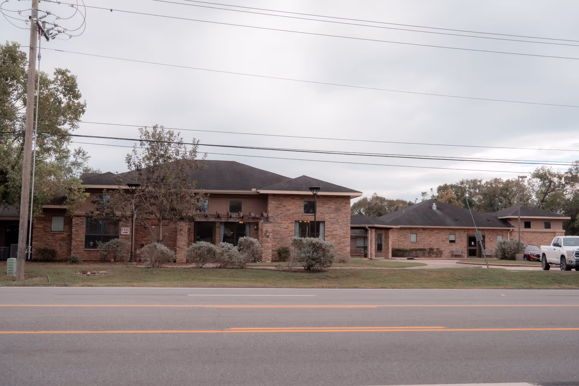 Front view of a low-rise brick senior living facility with a driveway and parked vehicles, seen from across the street under an overcast sky.