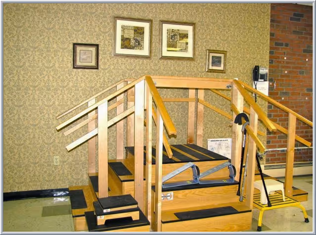 Indoor physical therapy stair-training apparatus with multiple wooden handrails, steps with nonslip treads, and a cane against a wallpapered wall with framed pictures.