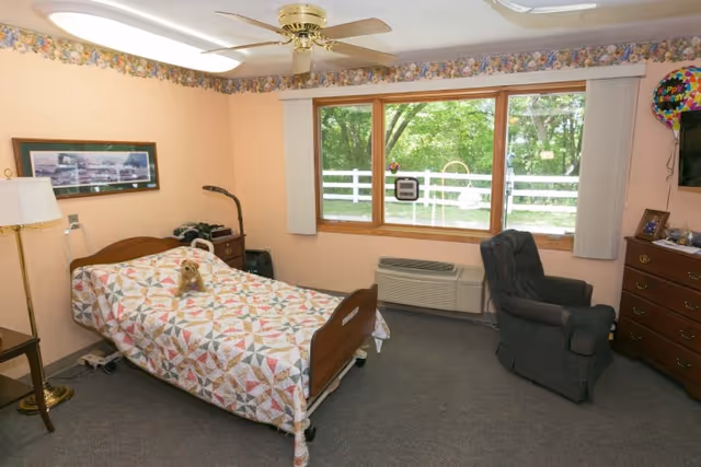 A cozy bedroom in a senior living facility with a single bed covered by a colorful quilt and a small stuffed animal on top. The room has a large window with vertical blinds showing a green outdoor view with a white fence. There is a dark upholstered armchair, a wooden dresser with a TV on top, and a floor lamp beside the bed. The walls are painted light peach with a floral border near the ceiling, and a ceiling fan is mounted above.